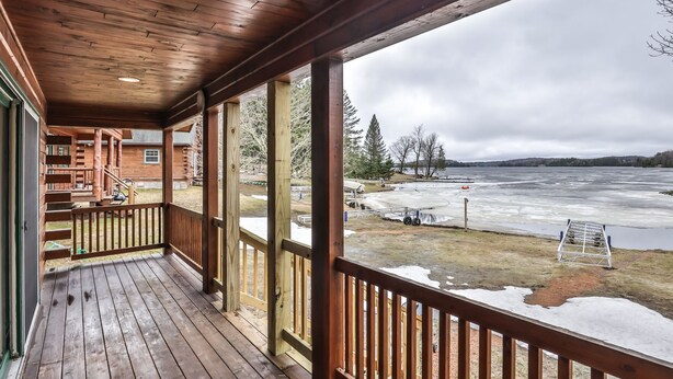 A wooden cabin with a porch overlooking a frozen lake, featuring a staircase and a view of the surrounding landscape. - Lakeside wilderness resort cabin with dock, fire pit, on-site store, bait shop! (Watersmeet)