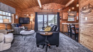 A cozy cabin interior with a living area featuring a sofa, armchairs, and a coffee table. There is a television, a fireplace, and a view of the outdoors through large windows. - Lakeside wilderness resort cabin with dock, fire pit, on-site store, bait shop! (Watersmeet)