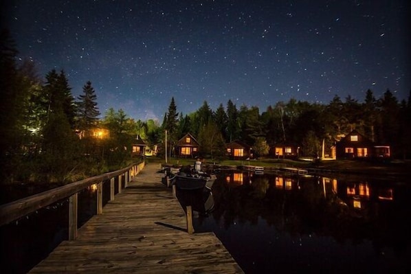A wooden dock extending into a calm body of water with houses on the opposite shore, under a starry night sky. - Lakeside wilderness resort cabin with dock, fire pit, on-site store, bait shop! (Watersmeet)