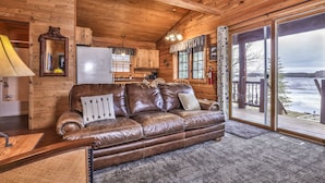 A cabin interior with a leather sofa, wooden walls, a kitchen area, and a large sliding glass door leading to a deck with a view of a lake. - Lakeside wilderness resort cabin with dock, fire pit, on-site store, bait shop! (Watersmeet)
