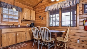 A wooden cabin interior with a kitchen area, a dining table with chairs, and a large window providing a view of the outdoors. - Lakeside wilderness resort cabin with dock, fire pit, on-site store, bait shop! (Watersmeet)