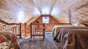 A cozy attic bedroom with a sloped ceiling, wooden beams, a bed, a chair, and a window. - Lakeside wilderness resort cabin with dock, fire pit, on-site store, bait shop! (Watersmeet)