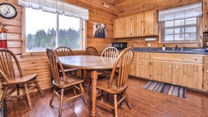 A wooden cabin interior with a dining table, chairs, a kitchen area, and a large window with a view of trees. - Lakeside wilderness resort cabin with dock, fire pit, on-site store, bait shop! (Watersmeet)