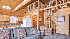 A cozy cabin interior with a wooden staircase, a sofa with patterned cushions, and a dining area with wooden cabinets and a refrigerator. - Lakeside wilderness resort cabin with dock, fire pit, on-site store, bait shop! (Watersmeet)