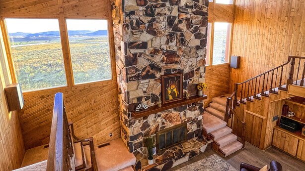 A spacious living area with a stone fireplace, wooden staircase, and large windows offering a view of the landscape. - Large cabin with amazing views, mountains, sunsets, and wildlife.
 (Pinedale)