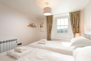 A hotel room with two beds, a radiator, a hanging light fixture, a window with curtains, and a coat rack with hangers.