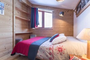 A cozy wooden room with a bed, a window with curtains, a lamp, and a shelf.