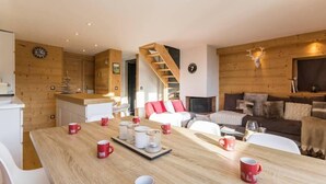 Accogliente interno di una baita con un tavolo da pranzo in legno apparecchiato per quattro persone, una scala, un caminetto e una zona living con un divano e un divanetto.