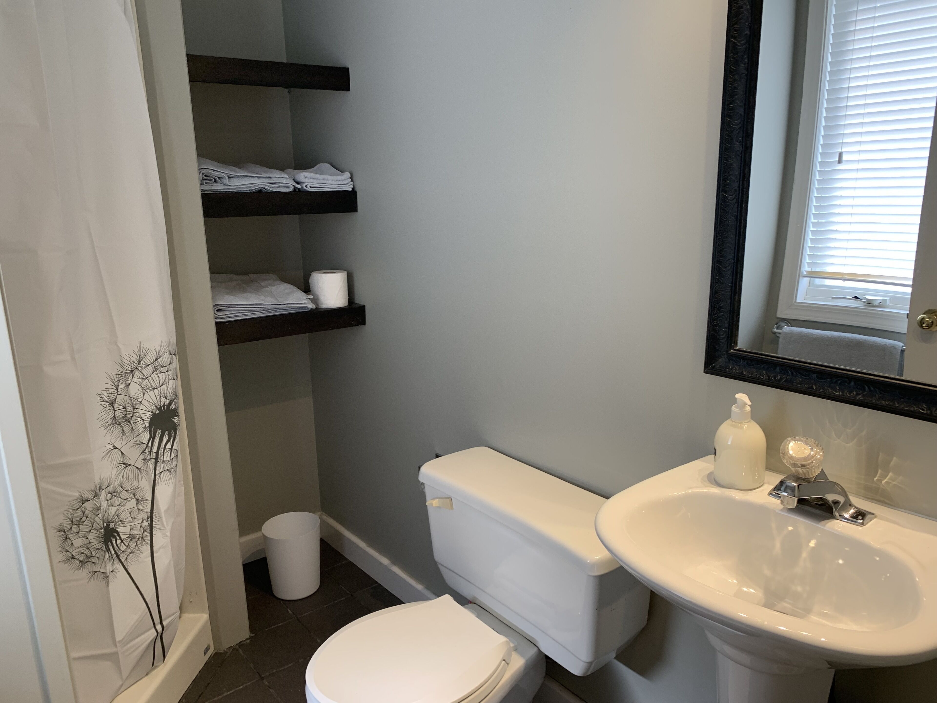 Un baño con inodoro, lavamanos, espejo y estantes con toallas y papel higiénico.