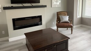 Ein Wohnzimmer mit Flachbildfernseher über einem Kamin, ein Holzsessel mit Kissen und ein kleiner Tisch.