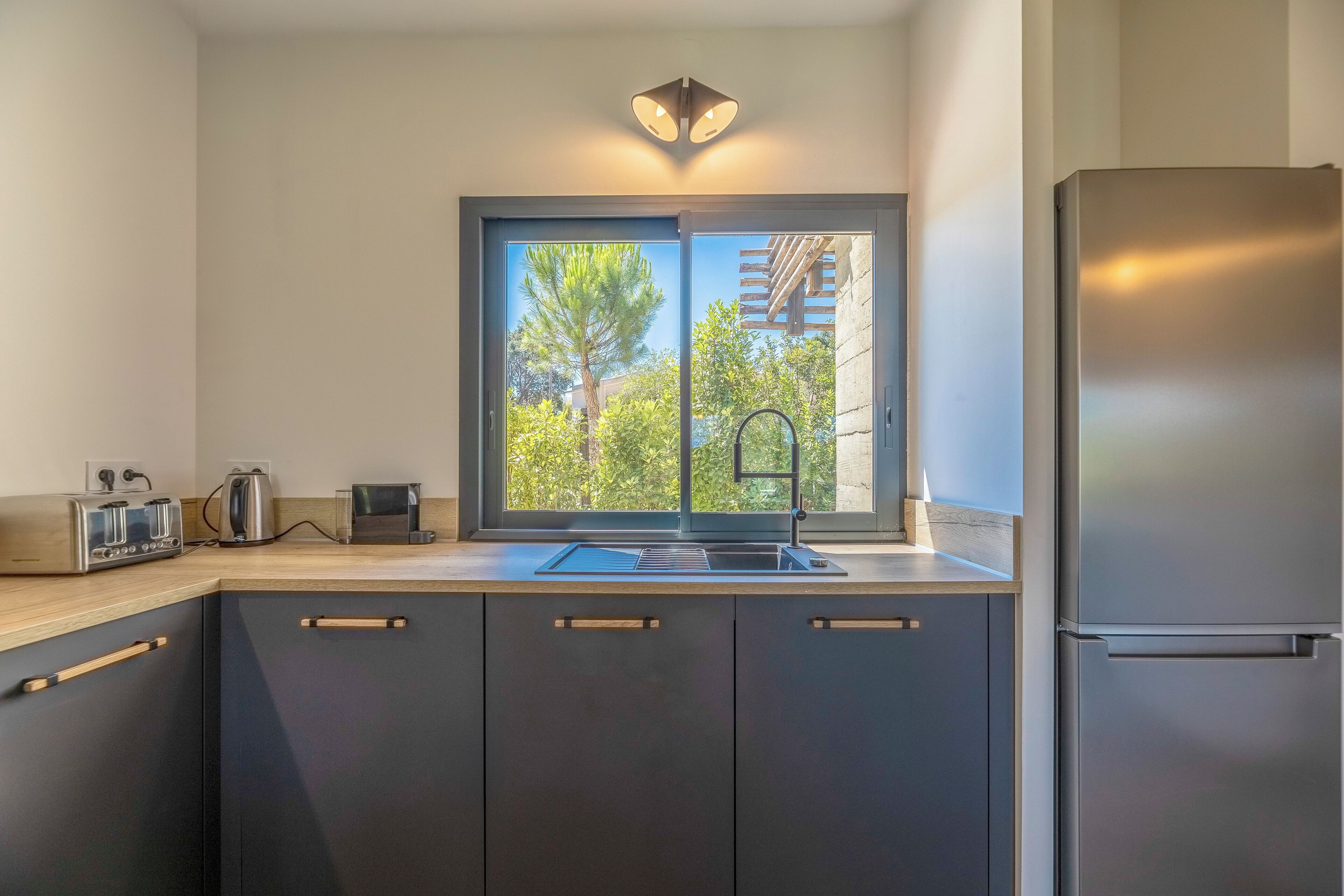 A modern kitchen with dark cabinets, a stainless steel refrigerator, a window with a view of trees, and a countertop with a toaster and kettle.
