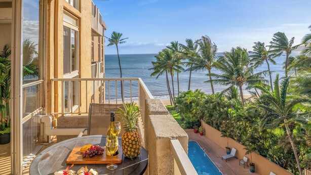 A balcony with a table set for two, overlooking a pool and palm trees, with a view of the ocean.