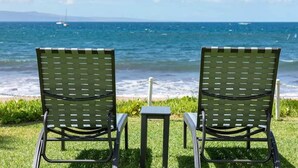 Two green metal chairs with a small table in between, facing a beach with the ocean and a sailboat in the distance.