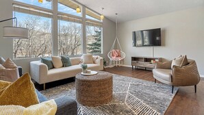 Un salon moderne avec un canapé, un pouf rond en osier, un fauteuil suspendu et un téléviseur à écran plat.