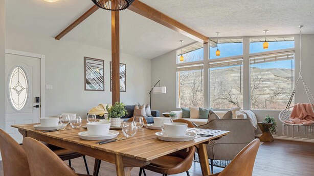 Un coin repas avec une table en bois dressée pour quatre personnes, entourée de fauteuils en cuir, et offrant une vue sur un paysage montagneux grâce à de grandes fenêtres.