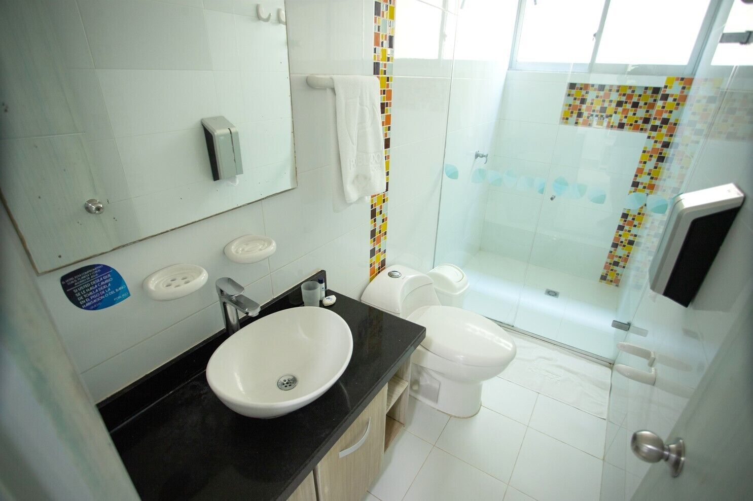 A bathroom with a white sink, toilet, and shower. There is a soap dispenser on the wall and a towel hanging on a rack.
