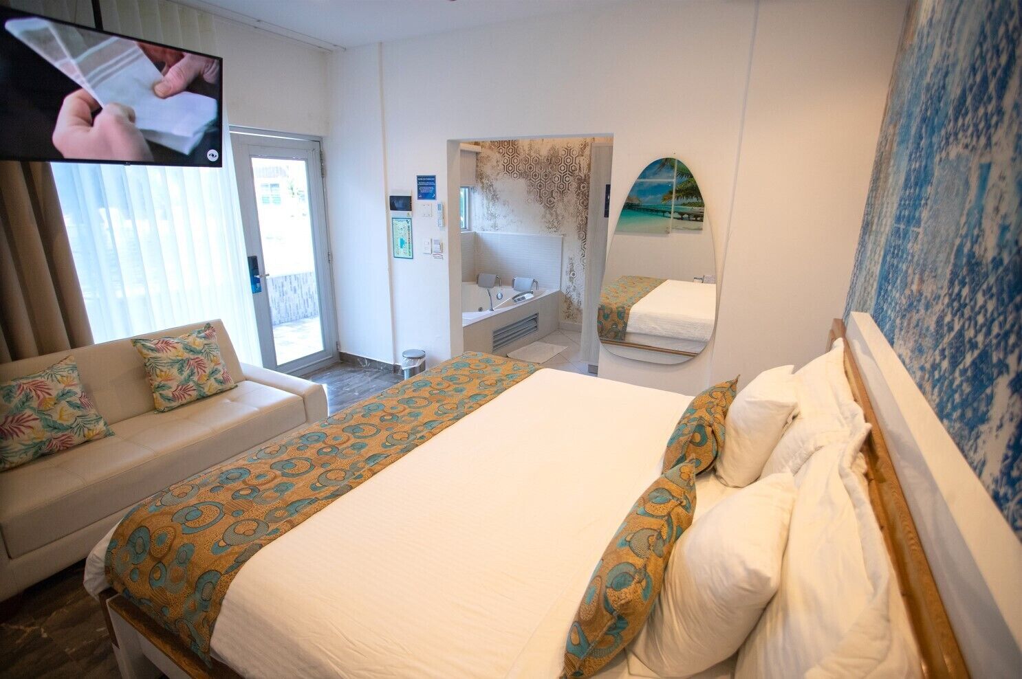 A hotel room with a bed, a sofa, a TV, a bathroom, and a view of the beach.