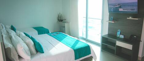 A hotel room with a bed, a desk, a television, and a window with a view of the sea.
