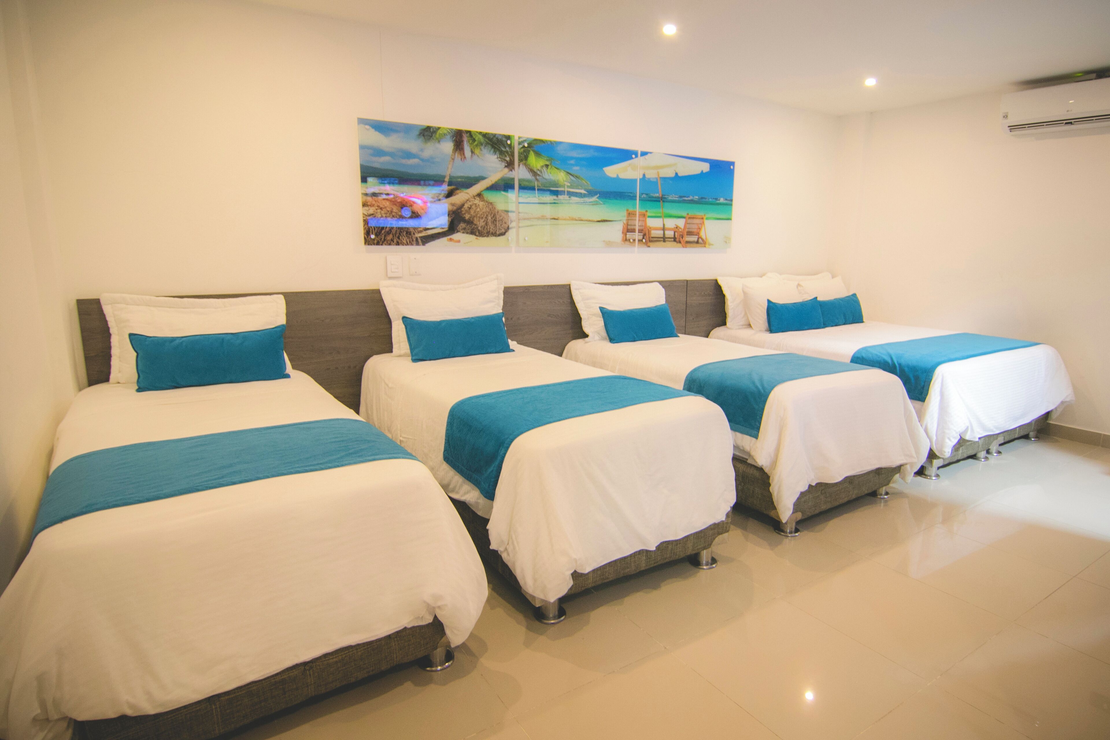Habitación de hotel con cuatro camas individuales, cada una con una colcha blanca y azul, y un cuadro grande con motivos playeros sobre las camas.