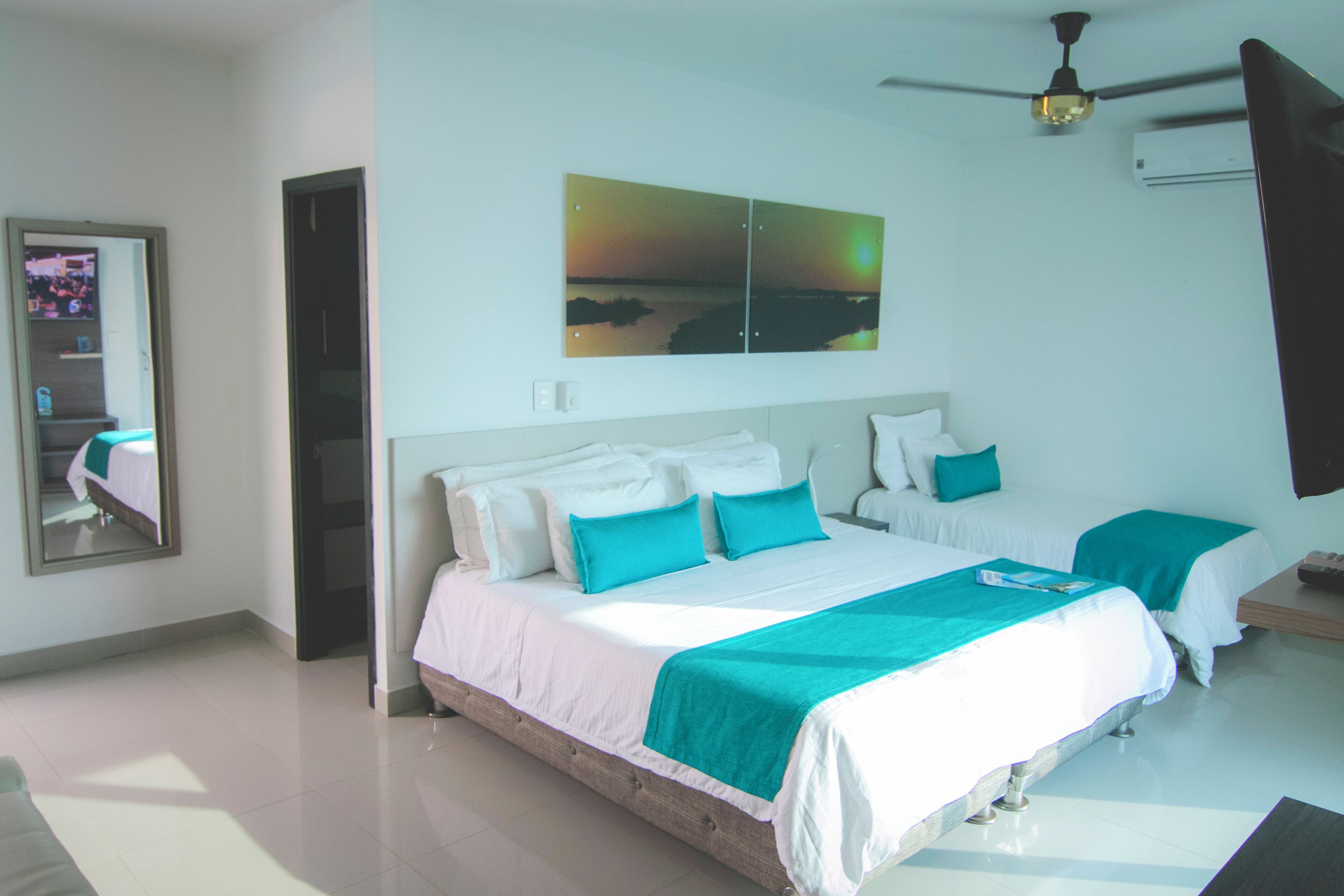 A hotel room with a large bed, a ceiling fan, and a painting of a sunset over water.