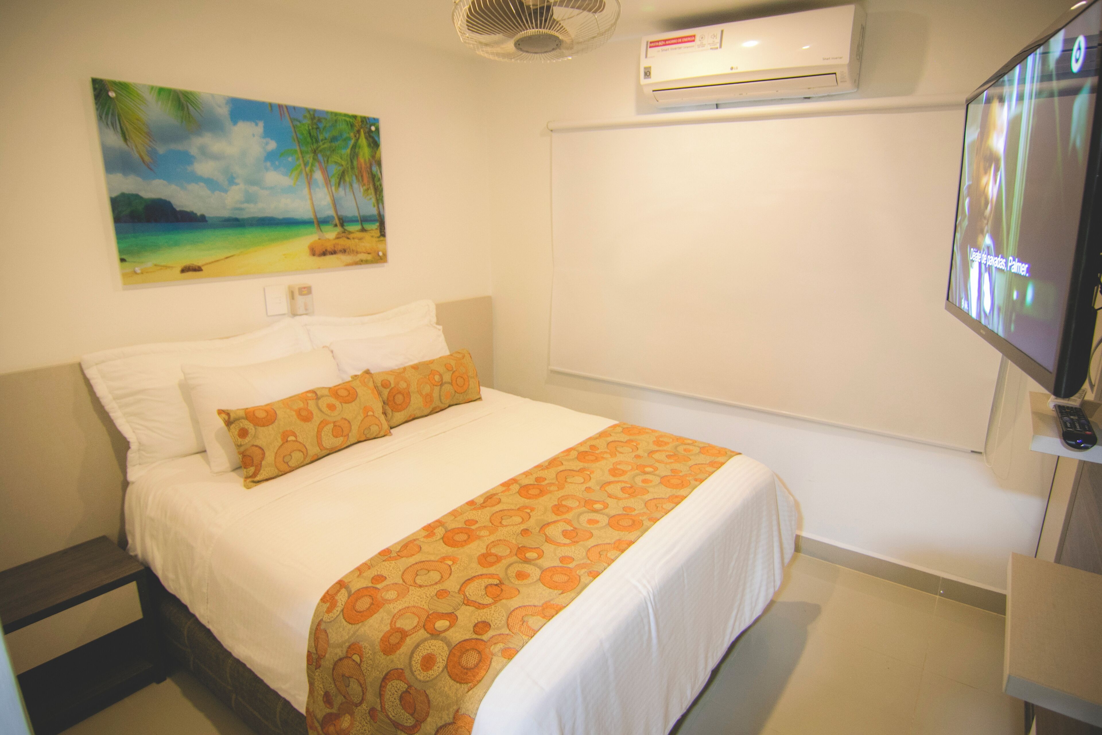 A hotel room with a bed, a painting of a beach, an air conditioner, and a television.