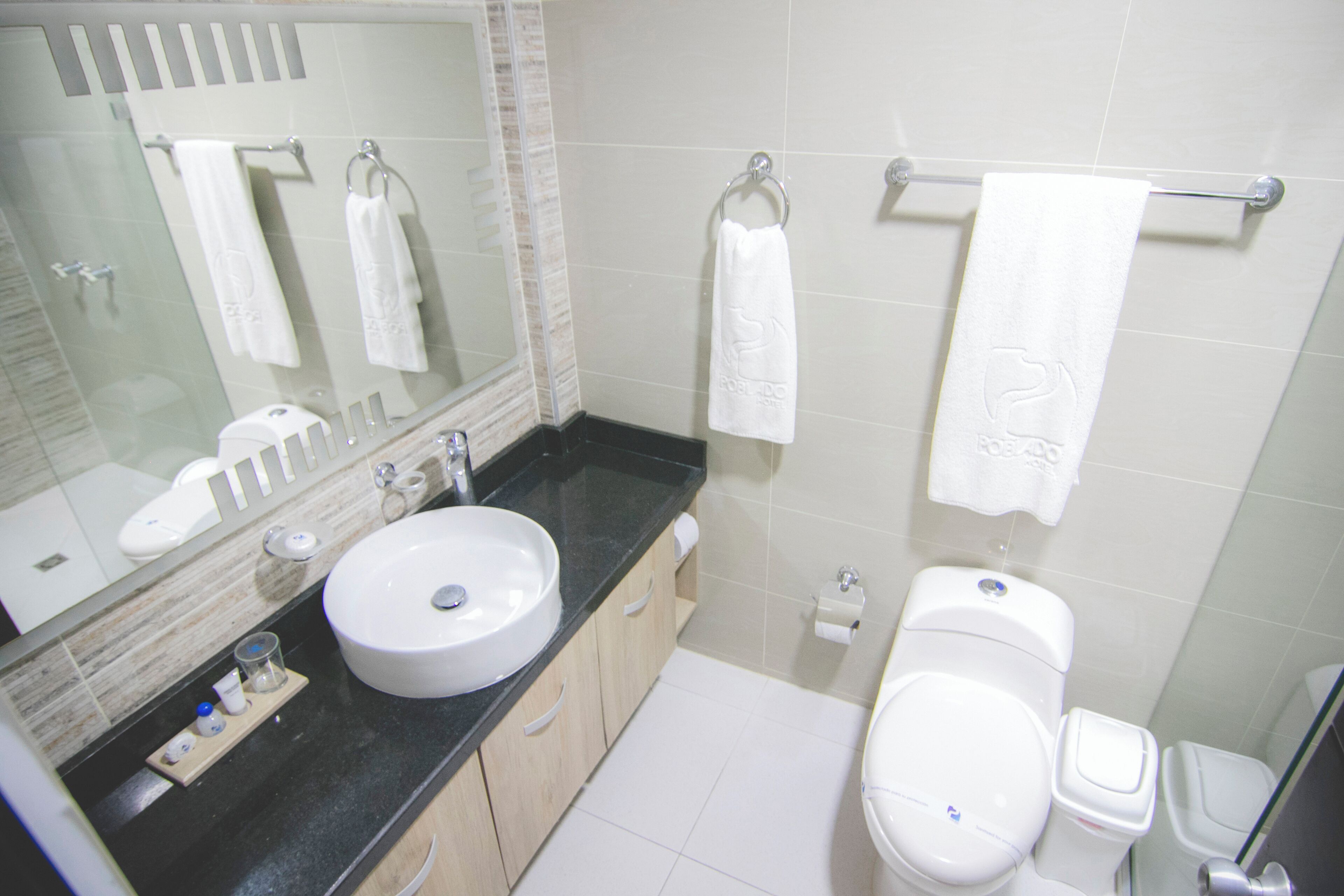 A modern bathroom with a large mirror, white sink, and toilet. There are multiple white towels hanging on towel bars.