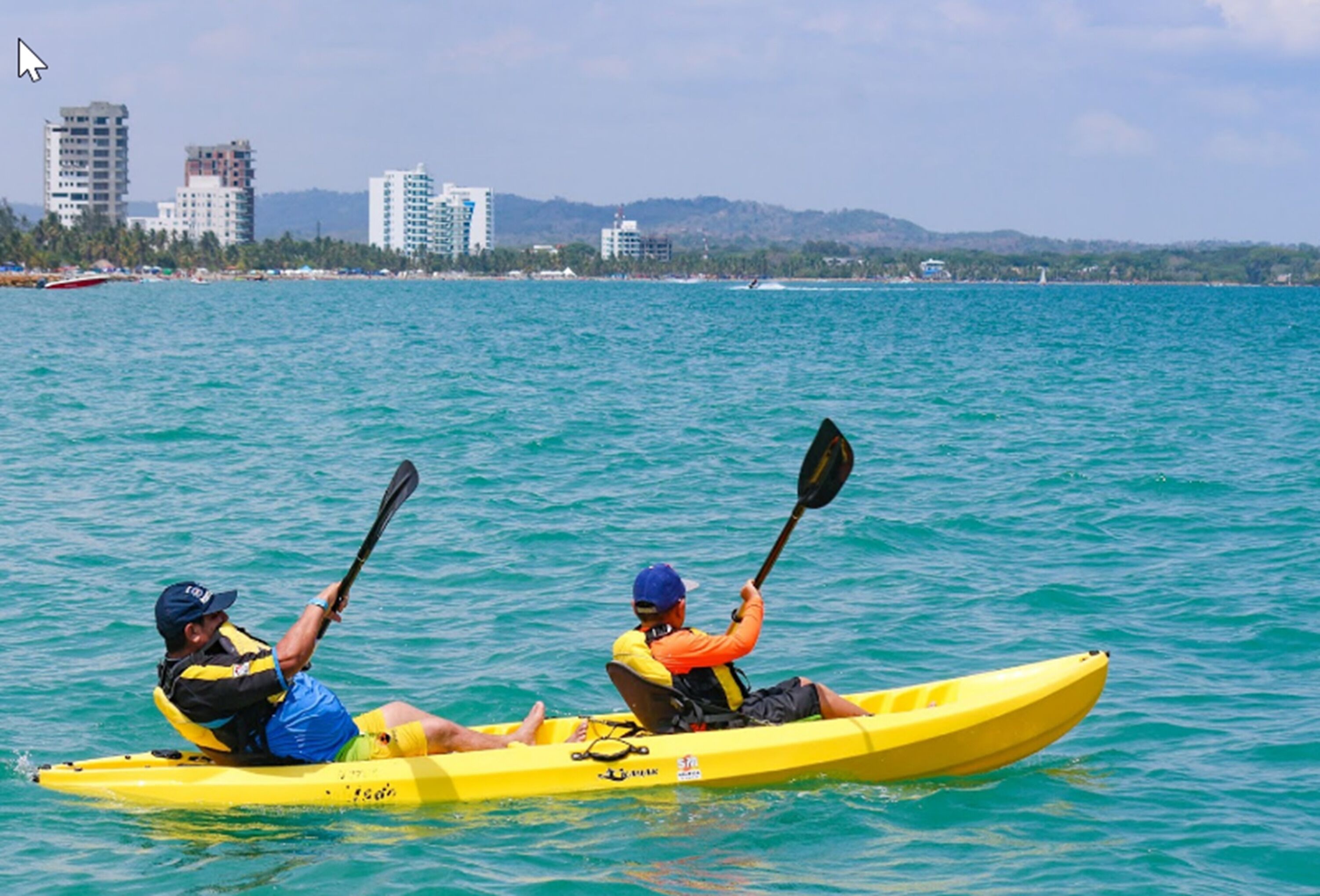 Deux personnes pratiquent le kayak sur une mer turquoise, à bord d’un kayak jaune, avec une vue sur la ville en arrière-plan.