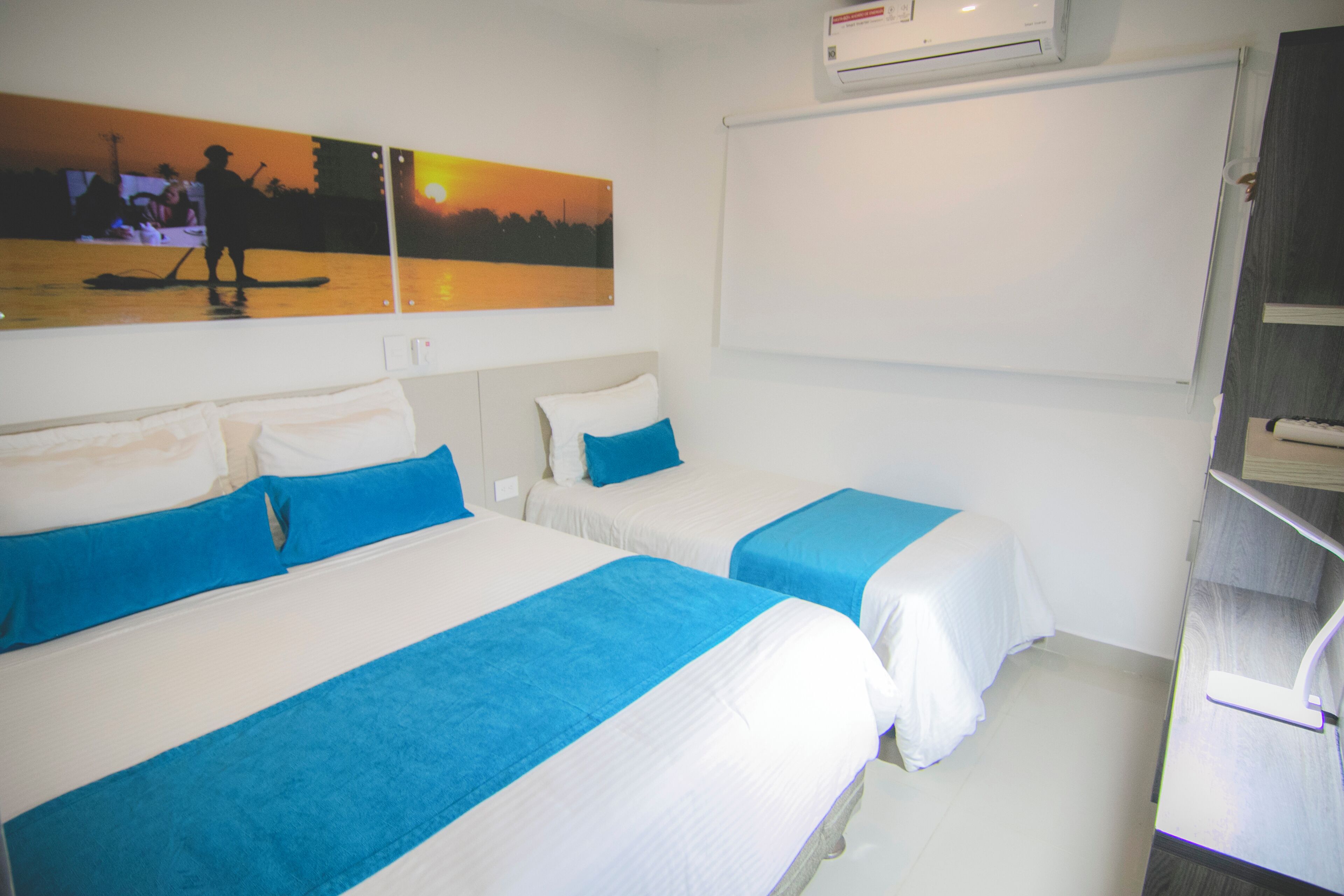A hotel room with two beds, each with blue and white bedding. There is a wall-mounted air conditioner and a painting depicting a sunset scene.