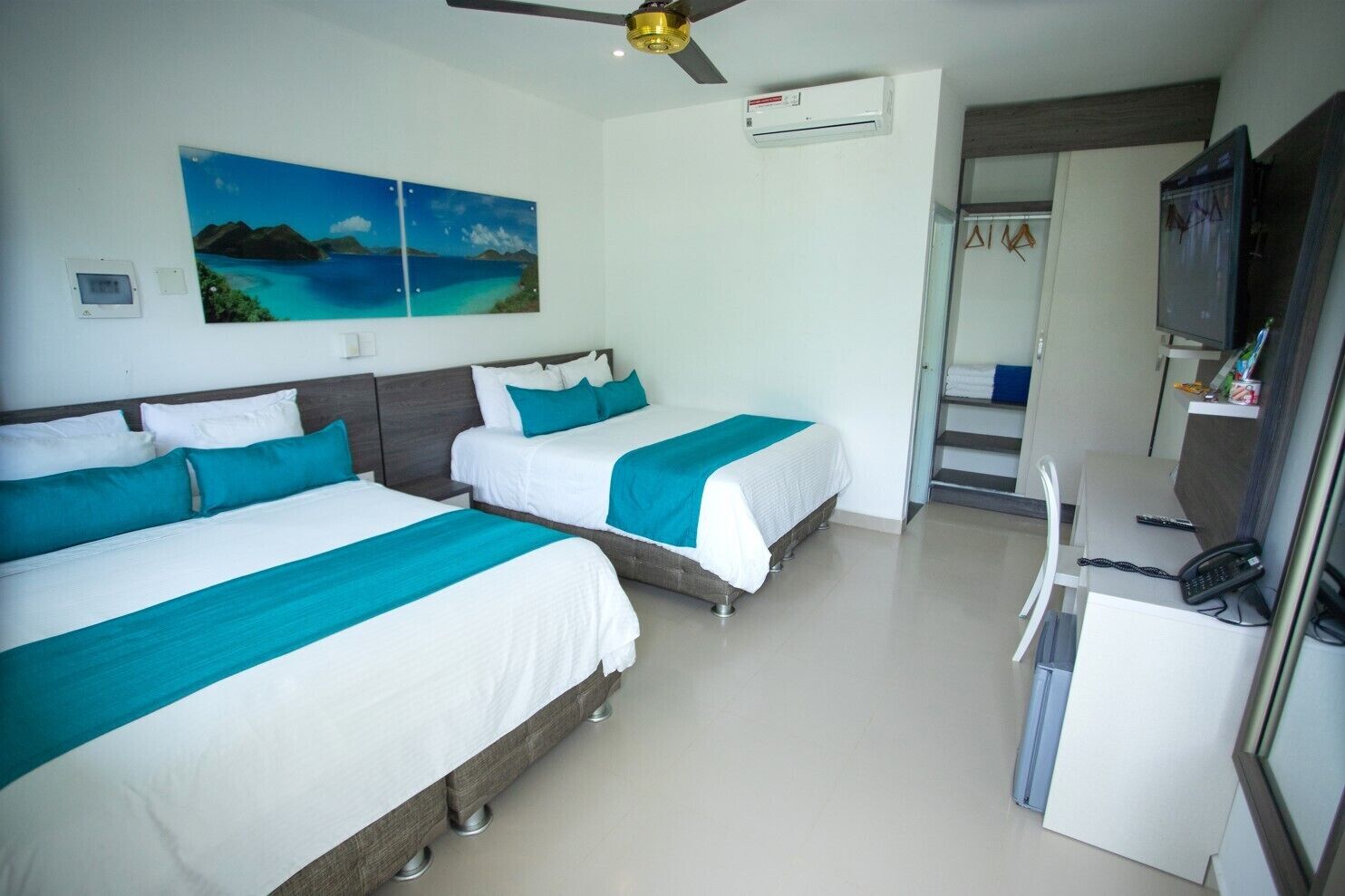 A hotel room with two beds, a desk, a chair, a TV, and a ceiling fan.