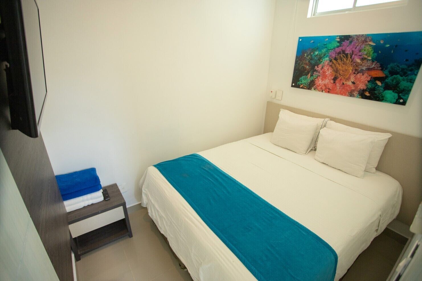 Un dormitorio con cama, mesita de noche, un cuadro de una escena submarina y ventana.