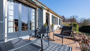 A patio with a table and chairs, a potted plant, and a view of a house with large windows and shutters. - Ellisa - Jolie Maison proche de la mer (Ver-sur-Mer)