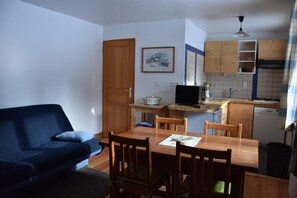 Un espacio habitable compacto con sofá, mesa de comedor, sillas, una pequeña cocina con armarios de madera, televisor y un cuadro en la pared.