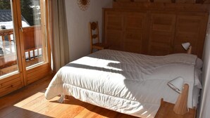 Une chambre avec une tête de lit en bois, un lit recouvert de linge de lit blanc, un plancher en bois, une fenêtre donnant sur un paysage enneigé et un balcon en bois.