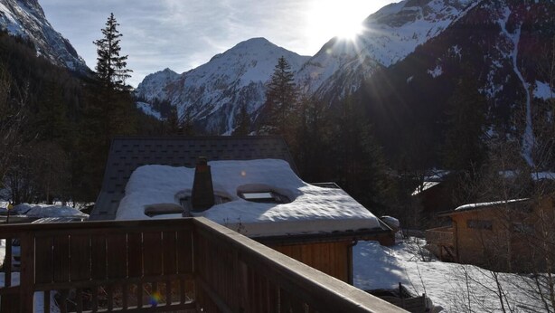 Une cabane en bois, dont le toit est recouvert de neige, entourée de montagnes et d’arbres.