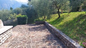 Un patio de piedra con pared de piedra y barandilla de metal, rodeado de follaje y árboles.