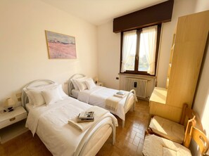 Une chambre d’hôtel avec deux lits, une fenêtre avec des rideaux, un radiateur, une chaise et un tableau au mur.