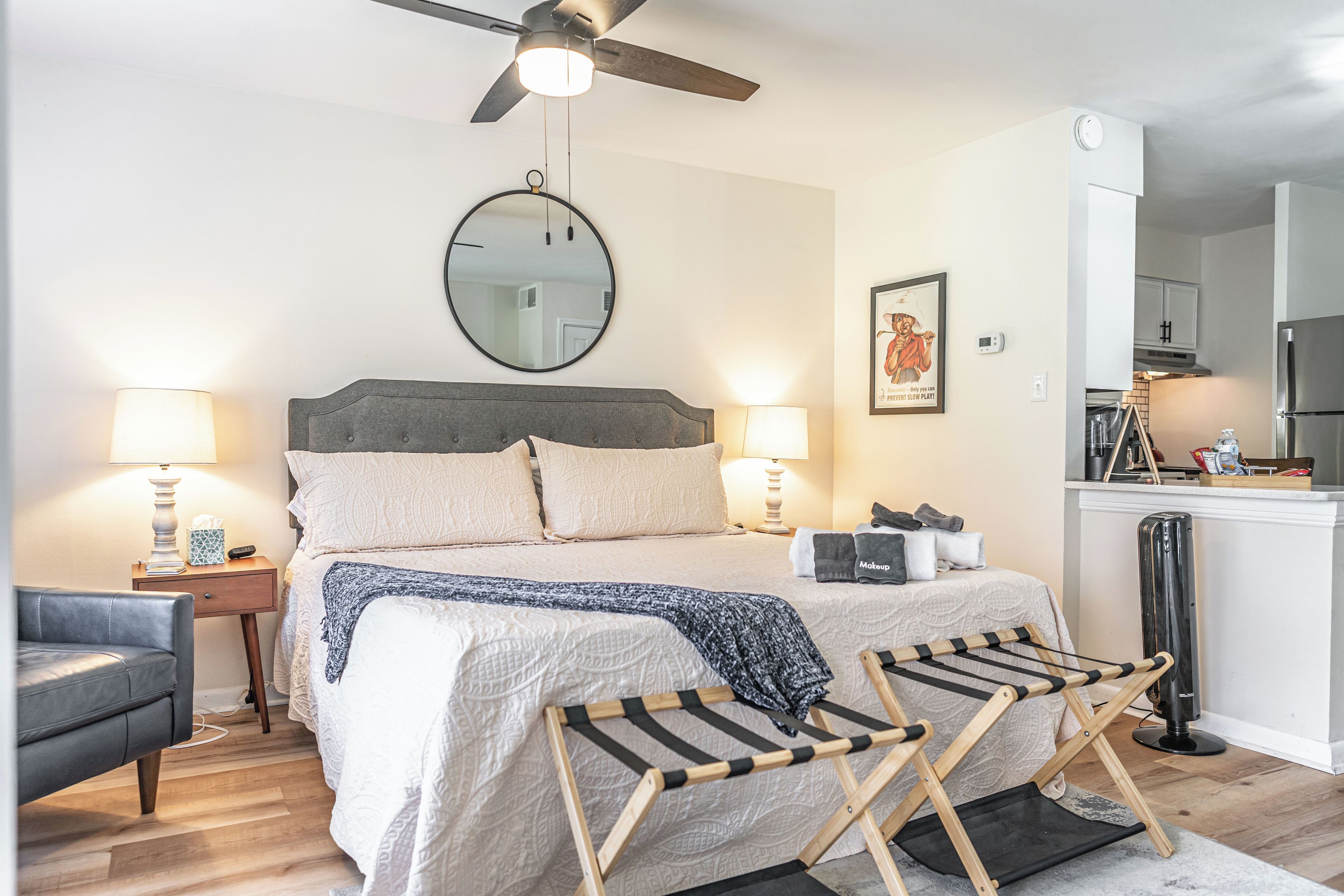 A bedroom with a bed, bedside lamps, a ceiling fan, a round mirror, a framed picture, and a view into the kitchen.