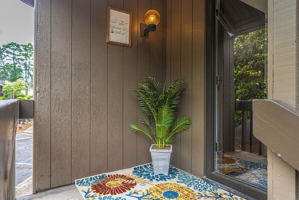 A wooden exterior wall with a potted plant, a framed sign, and a patterned mat at the entrance.