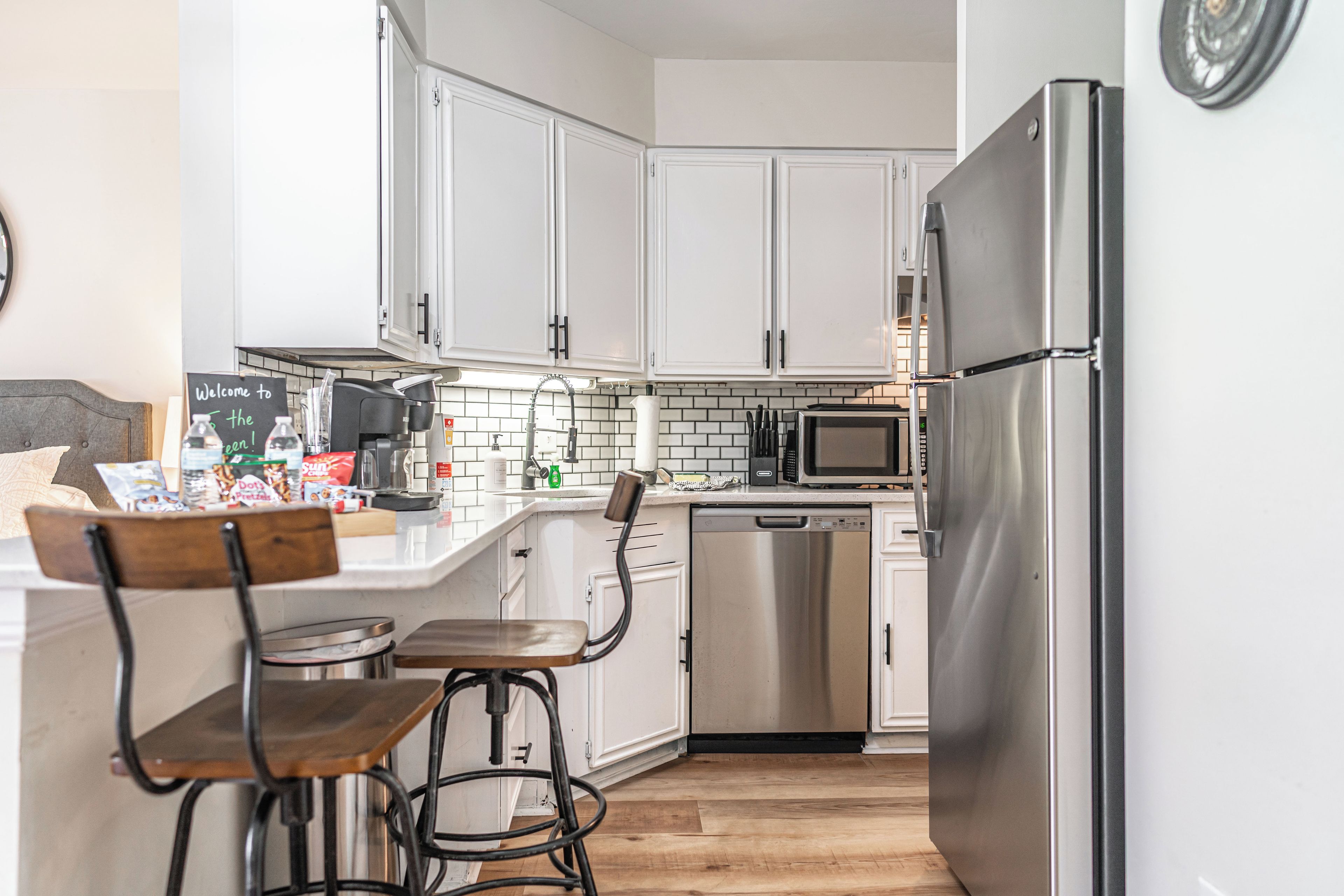 A modern kitchen with white cabinets, a stainless steel refrigerator, a dishwasher, and a microwave. There are two bar stools and a breakfast bar with various items on it.