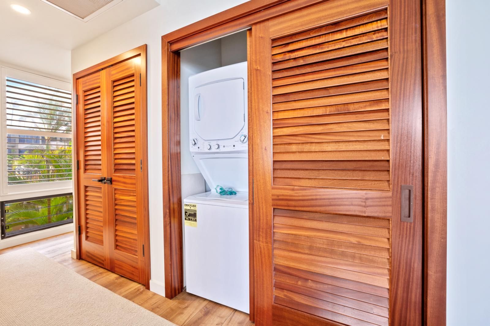 A laundry room with a front-loading washing machine, wooden doors, and a view of greenery outside.