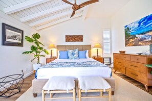 Ein Schlafzimmer mit einem Bett, Nachttischen, einer Kommode, einem Deckenventilator, einer großen Pflanze und Bildern an den Wänden.