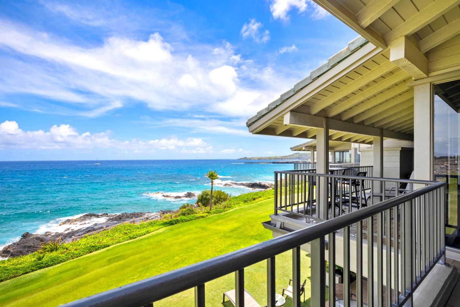 A balcony with a view of the ocean, a grassy area, and a building with a covered patio.