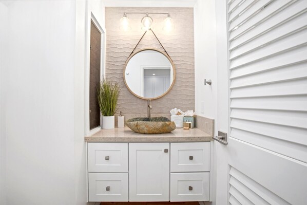 Une salle de bain avec un miroir rond, un lavabo en pierre et une plante en pot. Il y a deux armoires sous le miroir.