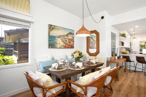 Un coin repas avec une table en bois dressée pour quatre personnes, entourée de chaises en osier. Un grand tableau représentant une scène de plage est accroché au-dessus de la table.