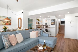 A modern living room with a blue sofa, a coffee table with wine glasses, and a kitchen in the background.