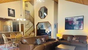 A spacious living room with a brown leather sofa, wooden dining table, and chairs. A staircase leads to an upper level.