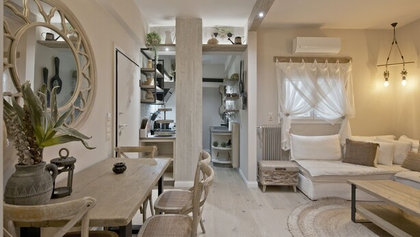 A cozy living room with a sofa, a dining table with chairs, a large mirror, and a kitchen area with shelves and a radiator. - Stylish Apartment at Pagrati (Vironas)