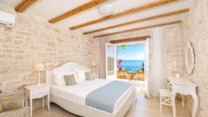 A bedroom with a stone wall, a bed, a nightstand, a bedside table, and a view of the sea. - The Yiameli Collection - Yasmi (Micro Nisi)
