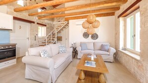 A cozy living room with two sofas, a wooden coffee table, and a stone wall. A staircase with wooden railings is visible in the background. - The Yiameli Collection - Yasmi (Micro Nisi)