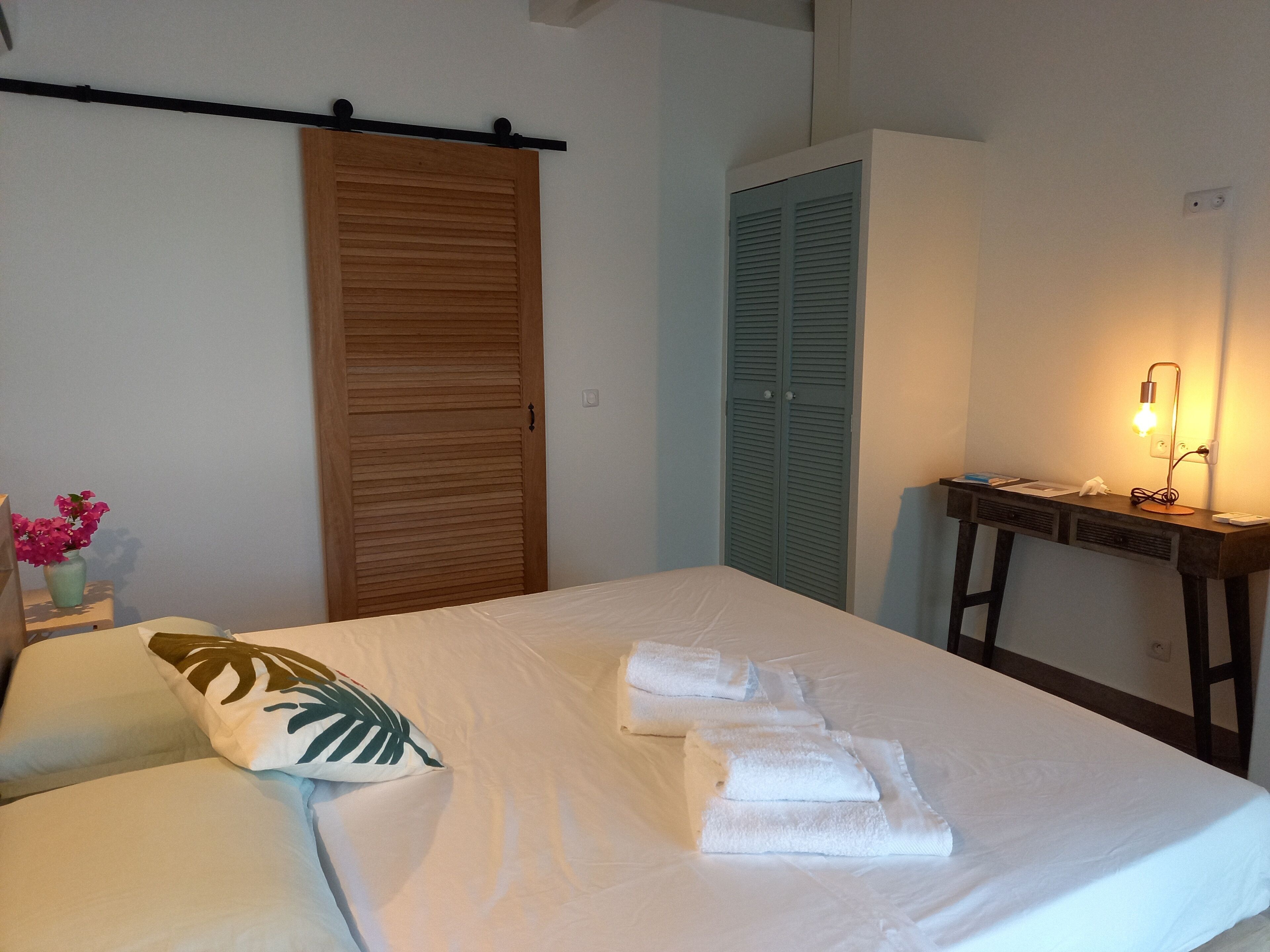 A neatly made bed with white linens and towels, a wooden wardrobe, a blue-clad closet, and a desk with a lamp.